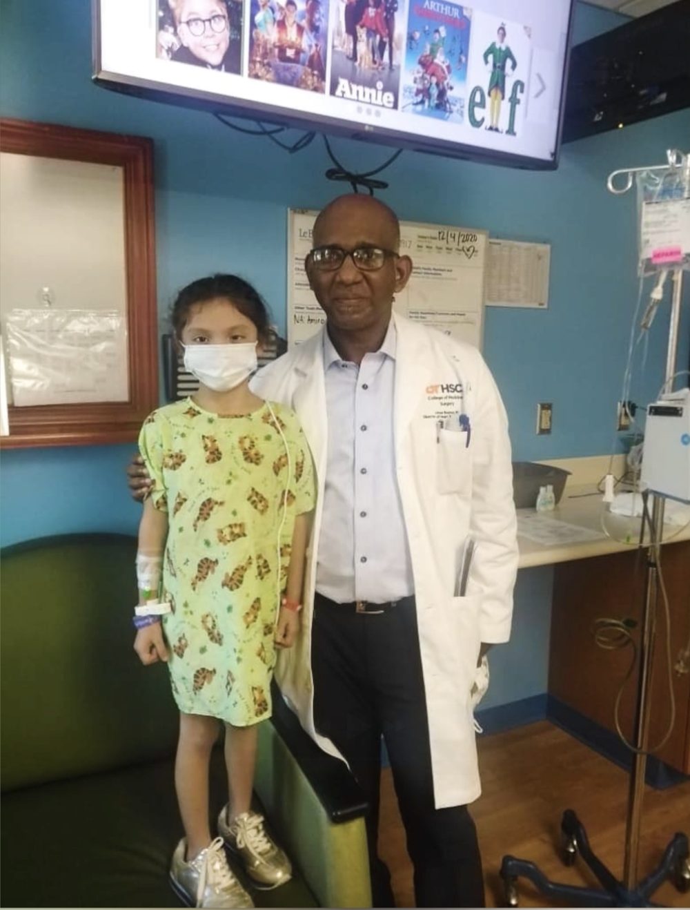 Valerie in a hospital gown and mask, standing on a chair beside her heart surgeon in a white coat.